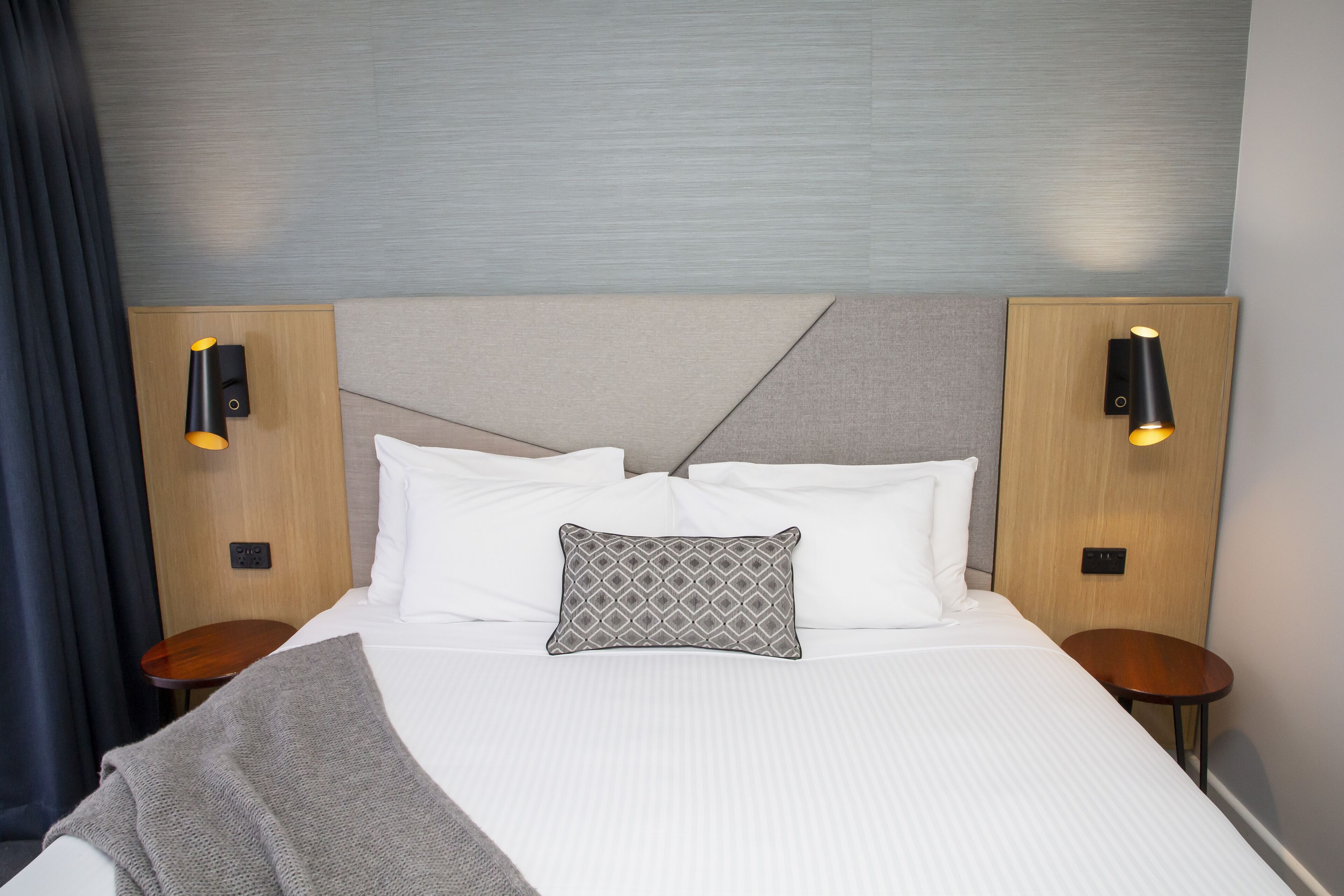 Ein ordentlich bezogenes Bett mit weißen Bettwäsche, einer grauen Decke und einem gemusterten Kissen. Es gibt zwei Nachttische mit Lampen, und das Zimmer hat ein modernes Design mit einem grauen Kopfteil und Holzelementen.