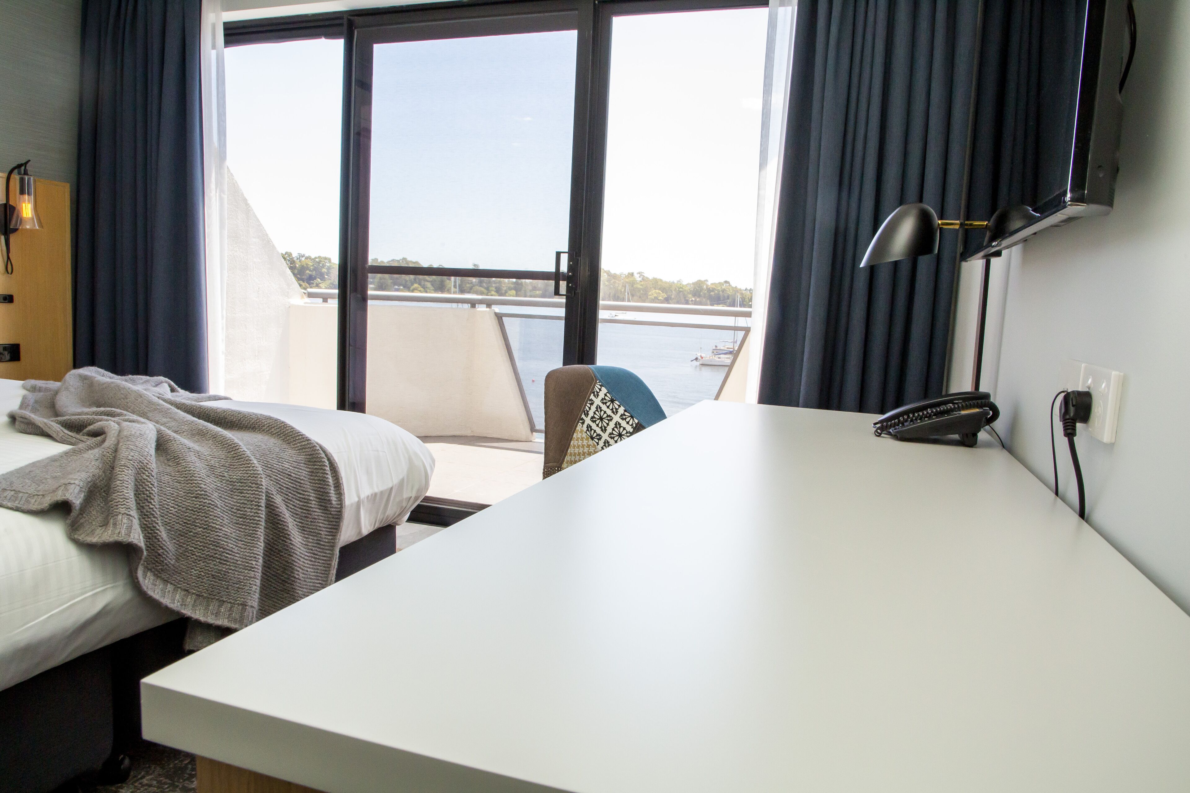 A hotel room with a bed, a desk, a chair, and a view of a body of water through large windows.