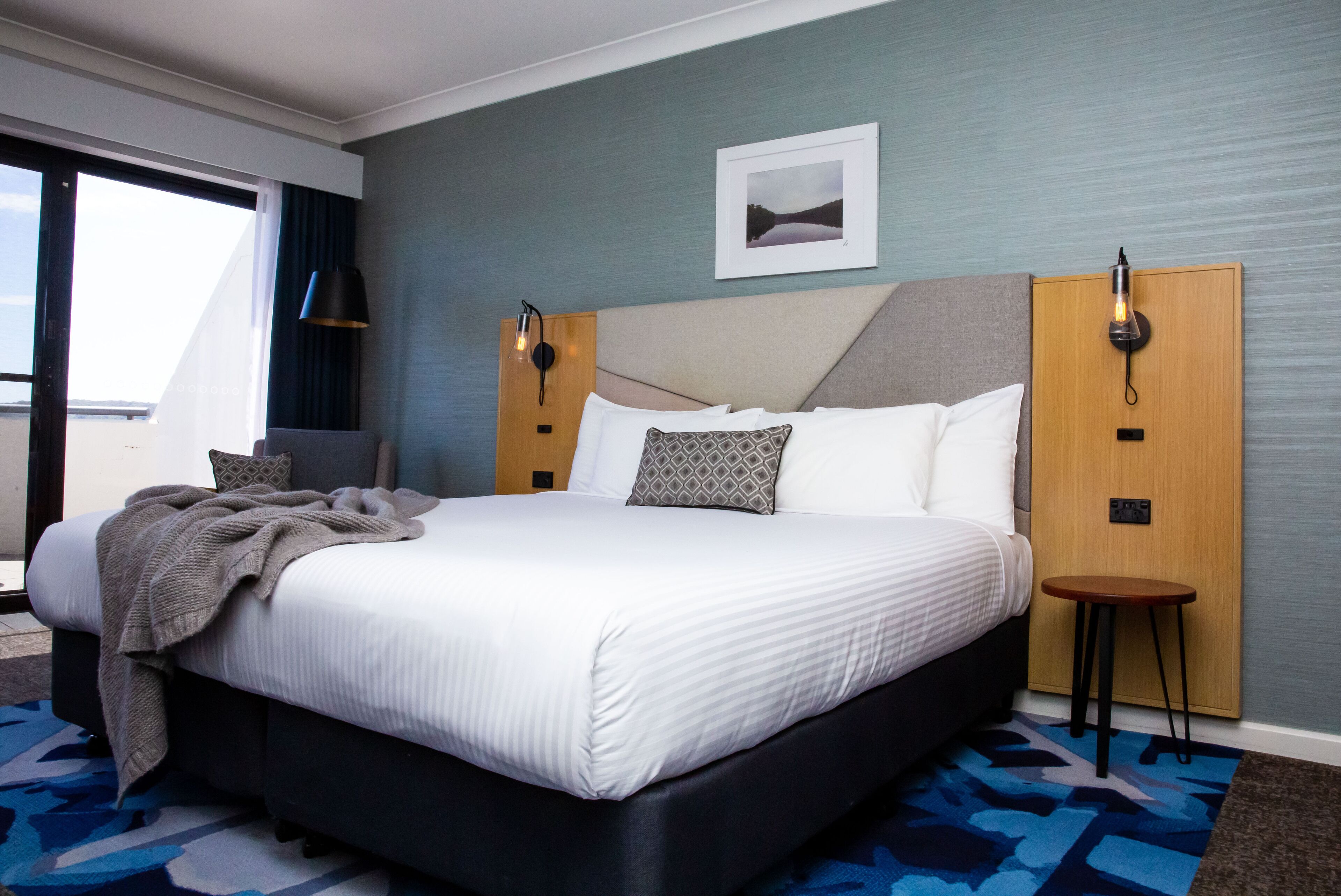 A hotel room with a large bed, a side table, a nightstand, a picture on the wall, and a window with curtains.