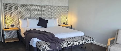 A hotel room with a large bed, a bedside table with a lamp, a chair, and a patterned headboard.