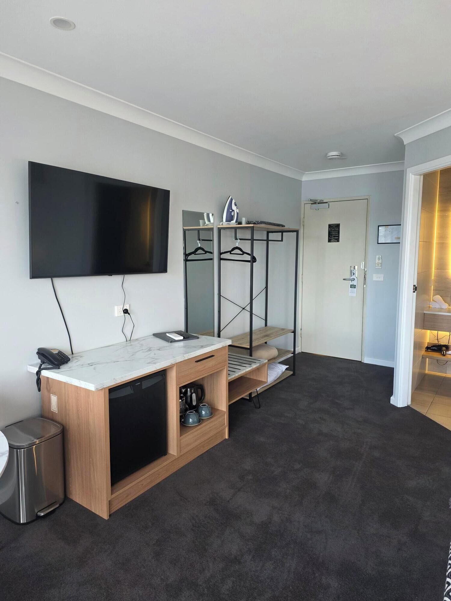 Una camera d'albergo moderna con TV a schermo piatto, una toeletta in legno con lavandino, un frigorifero compatto e un piccolo cestino.