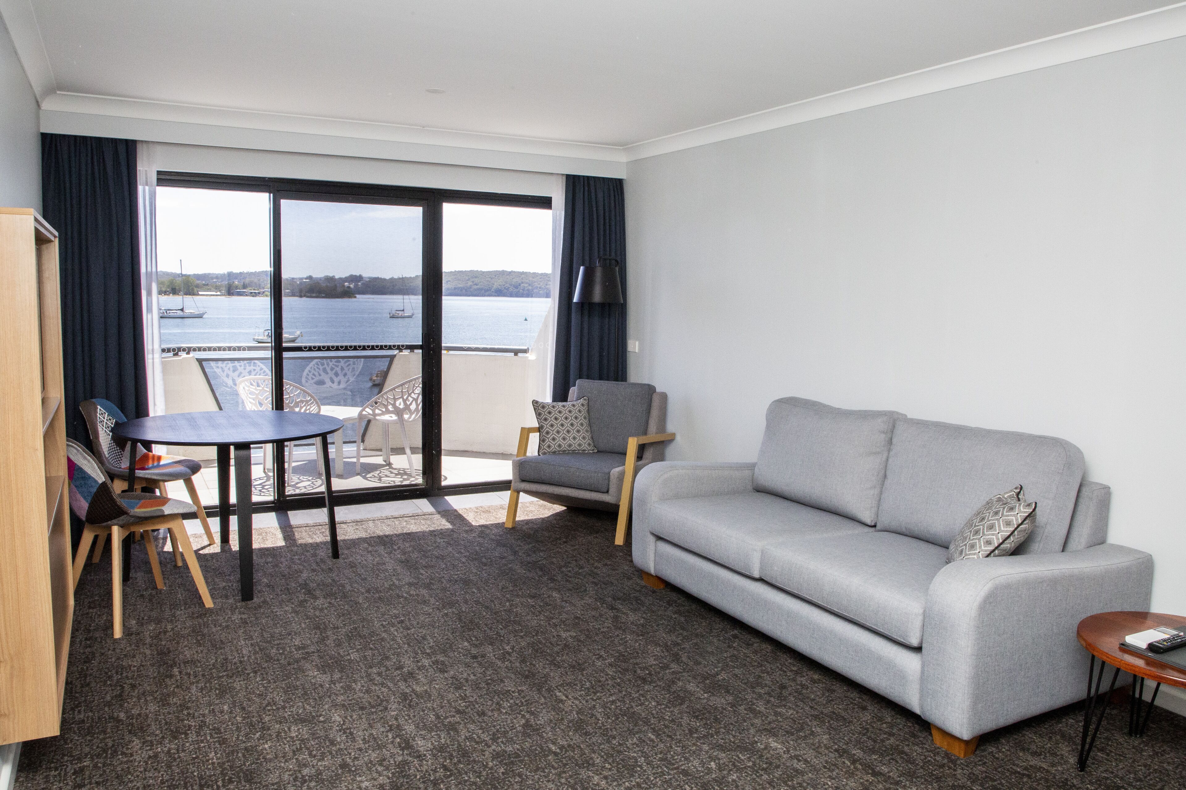 A living room with a grey sofa, a small round table, a chair, and a bookshelf. There is a large window with a view of the water and a balcony.