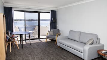 Un salon avec un canapé gris, une petite table ronde, un fauteuil et une bibliothèque. Il y a une grande fenêtre donnant sur l’eau et un balcon.