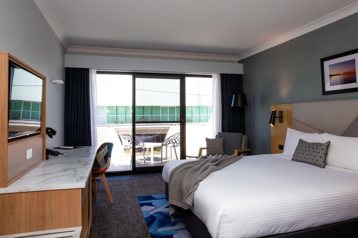 A hotel room with a bed, a desk with a chair, a desk lamp, a telephone, a window with curtains, and a view of the outside balcony.