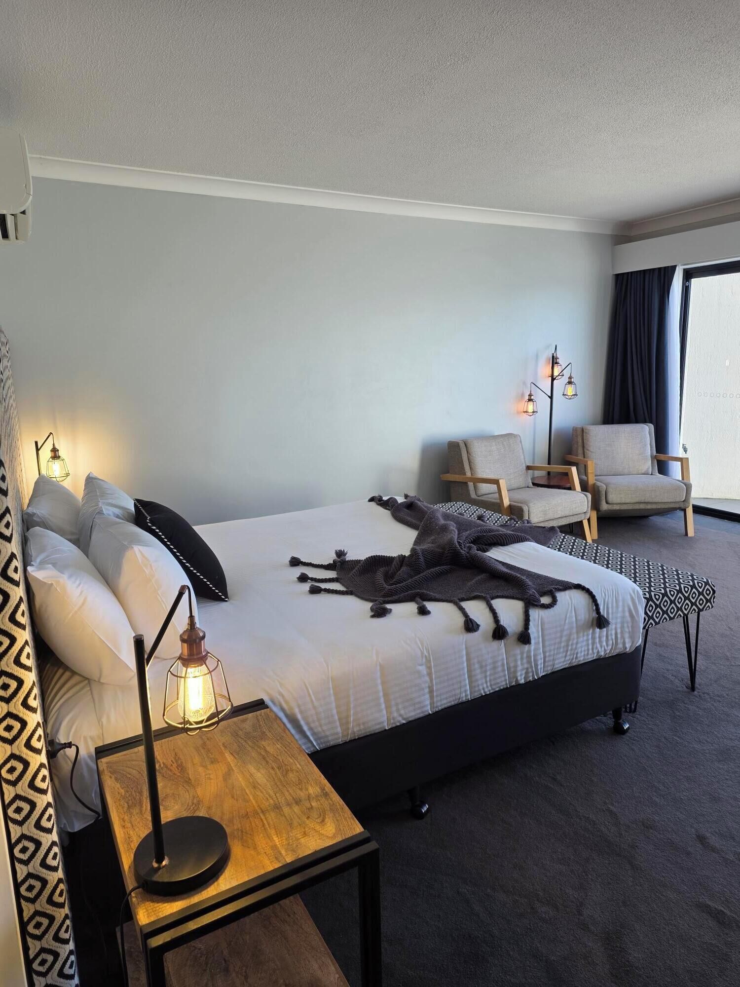 Quarto de hotel com cama, duas cadeiras, mesa de cabeceira com abajur e janela com cortinas.