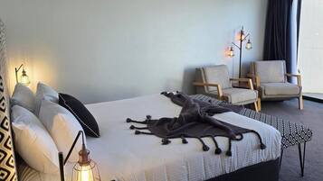 Quarto de hotel com cama, duas cadeiras, mesa de cabeceira com abajur e janela com cortinas.