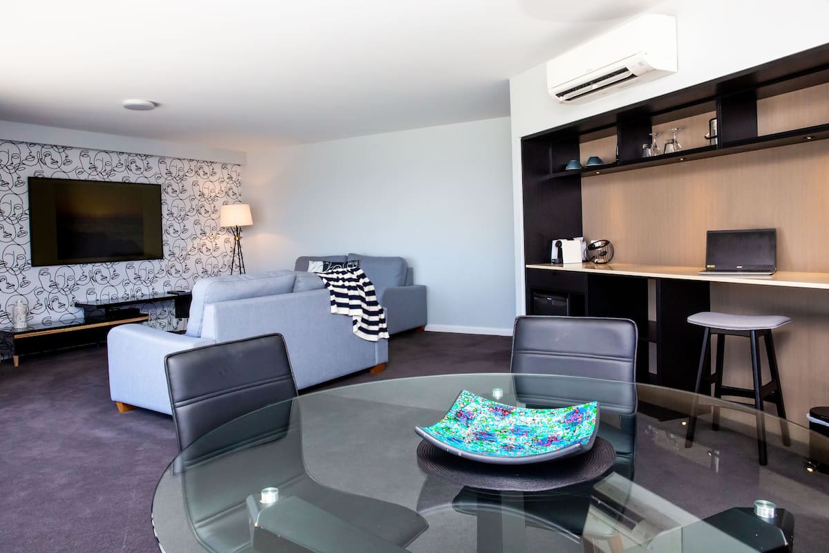 A modern living room with a glass dining table, a sofa, a TV, and a desk with a laptop.