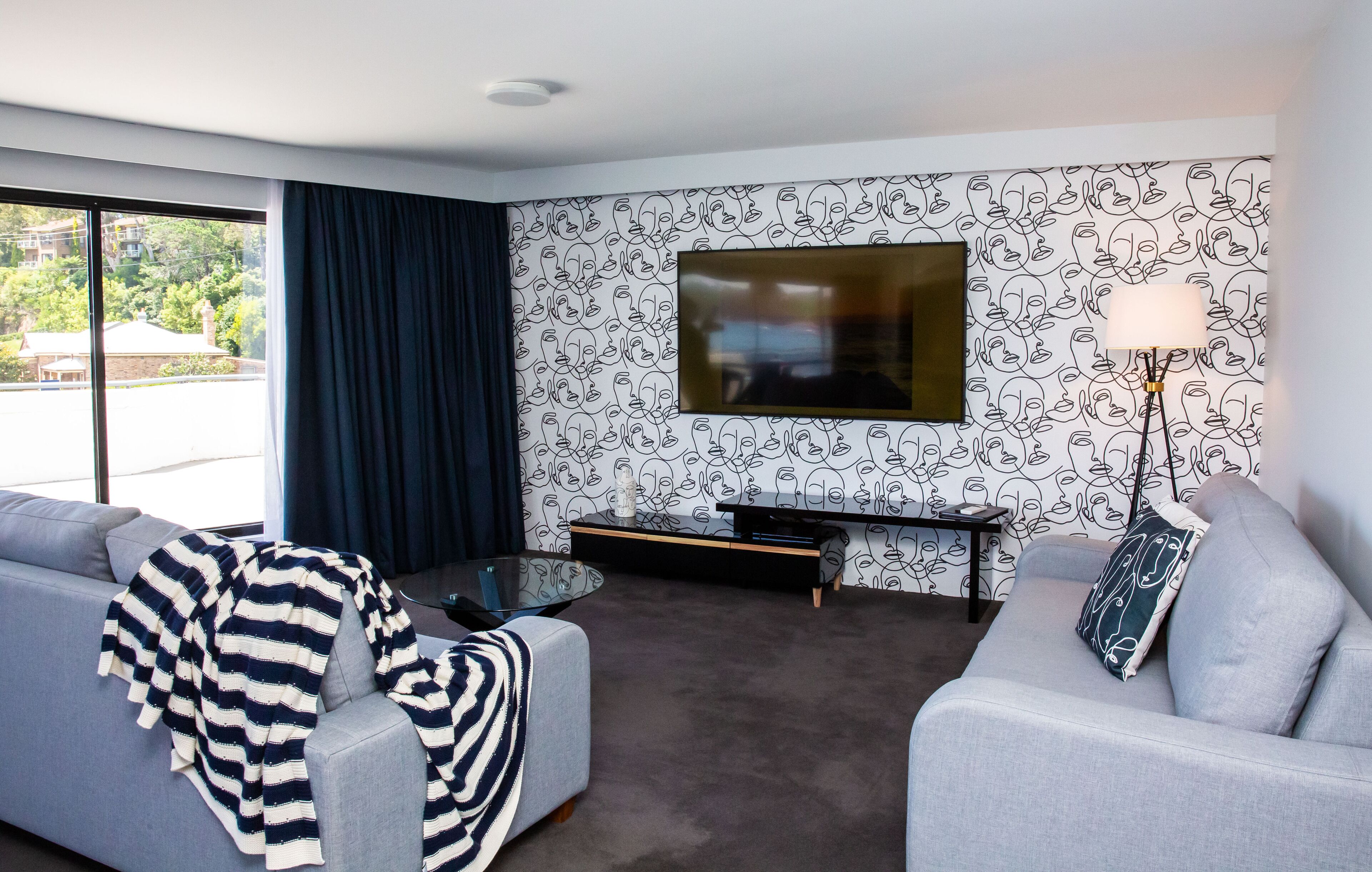 A modern living room with a grey sofa, a striped throw blanket, a coffee table, and a TV mounted on a wall with a face pattern wallpaper.