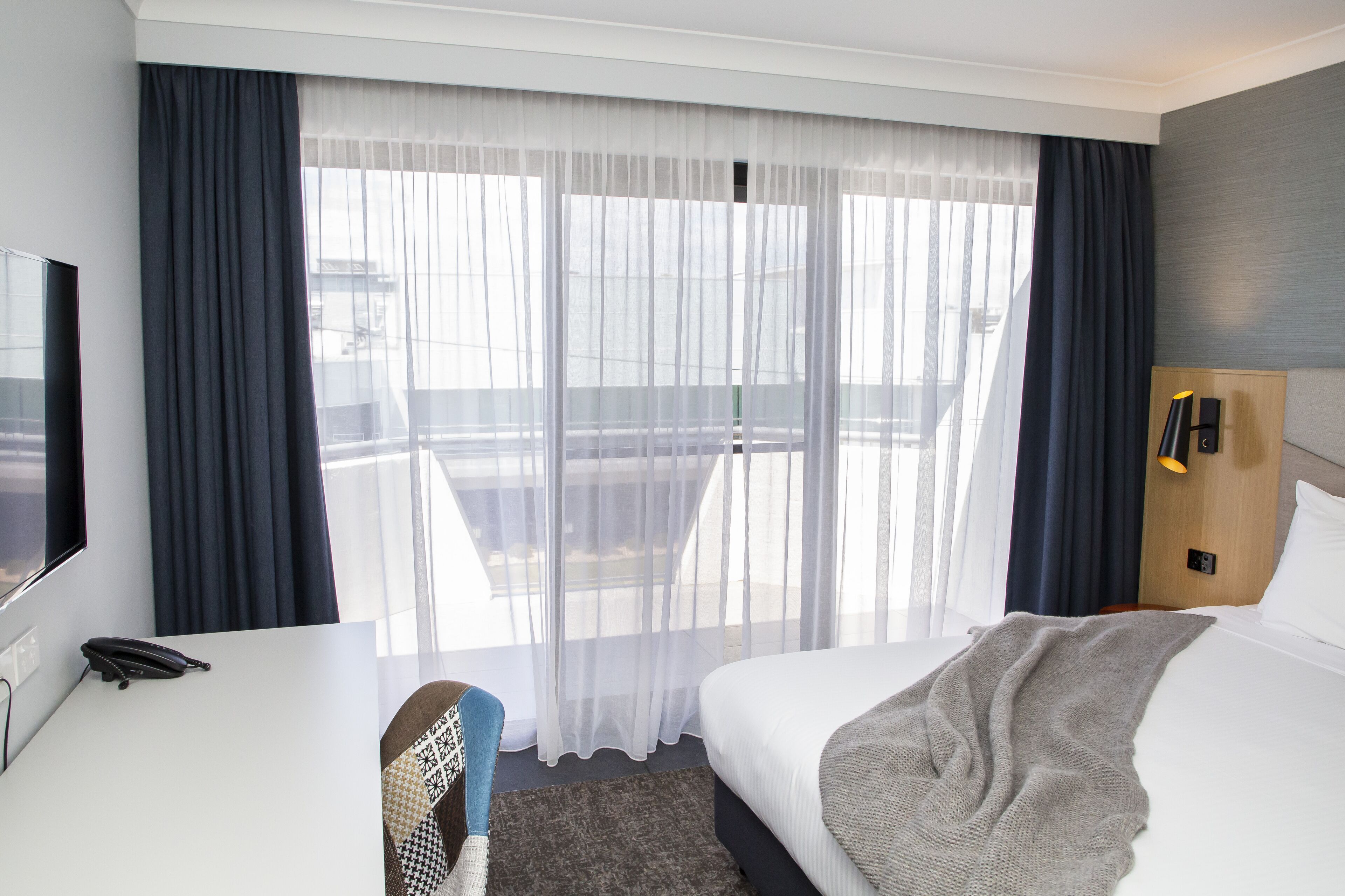 Habitación de hotel con cama, escritorio con teléfono, silla, televisión y ventana con cortinas.