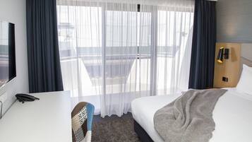 Habitación de hotel con cama, escritorio con teléfono, silla, televisión y ventana con cortinas.