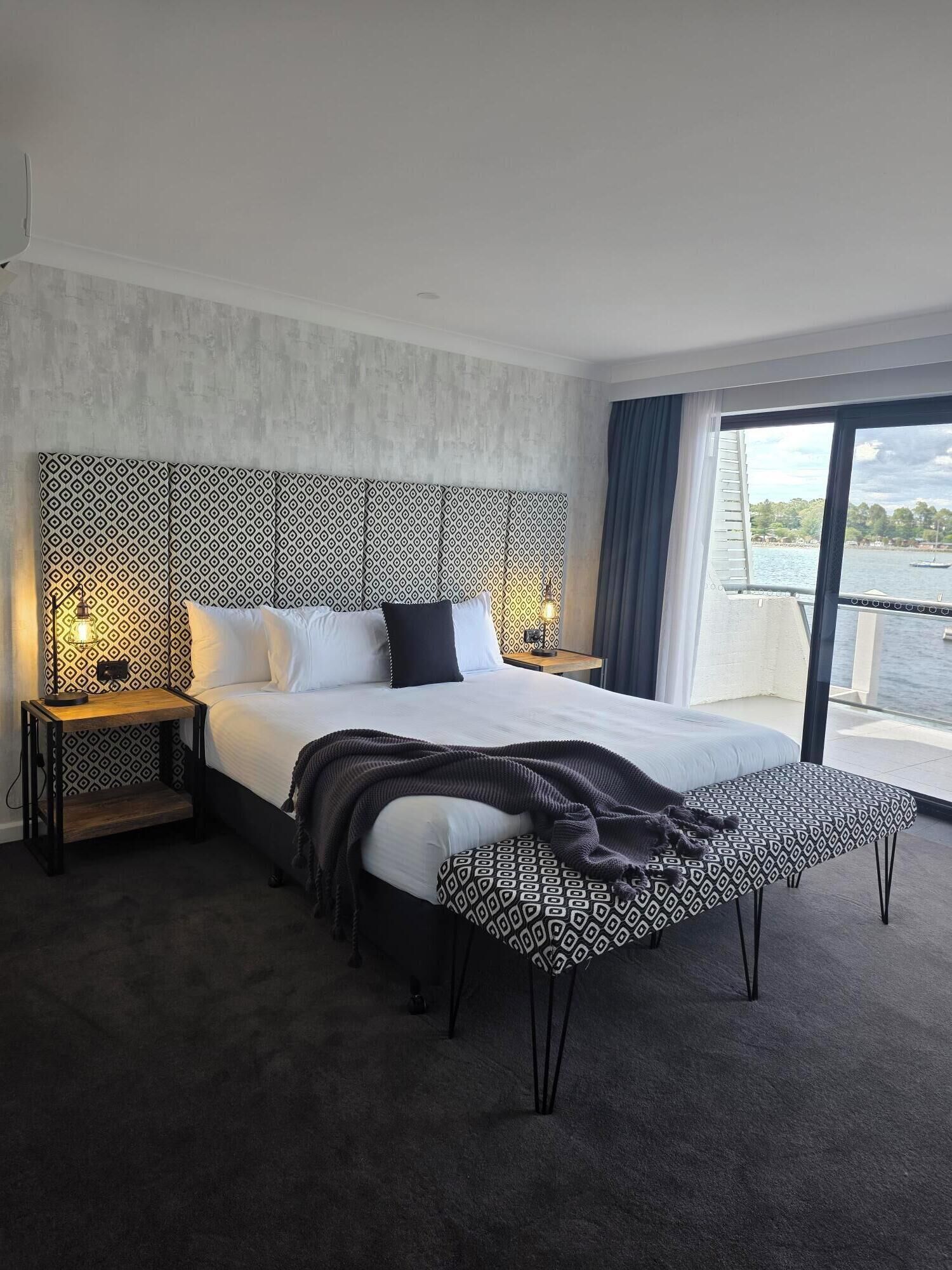 A modern hotel room with a large bed, two bedside tables with lamps, a view of a body of water through a balcony.