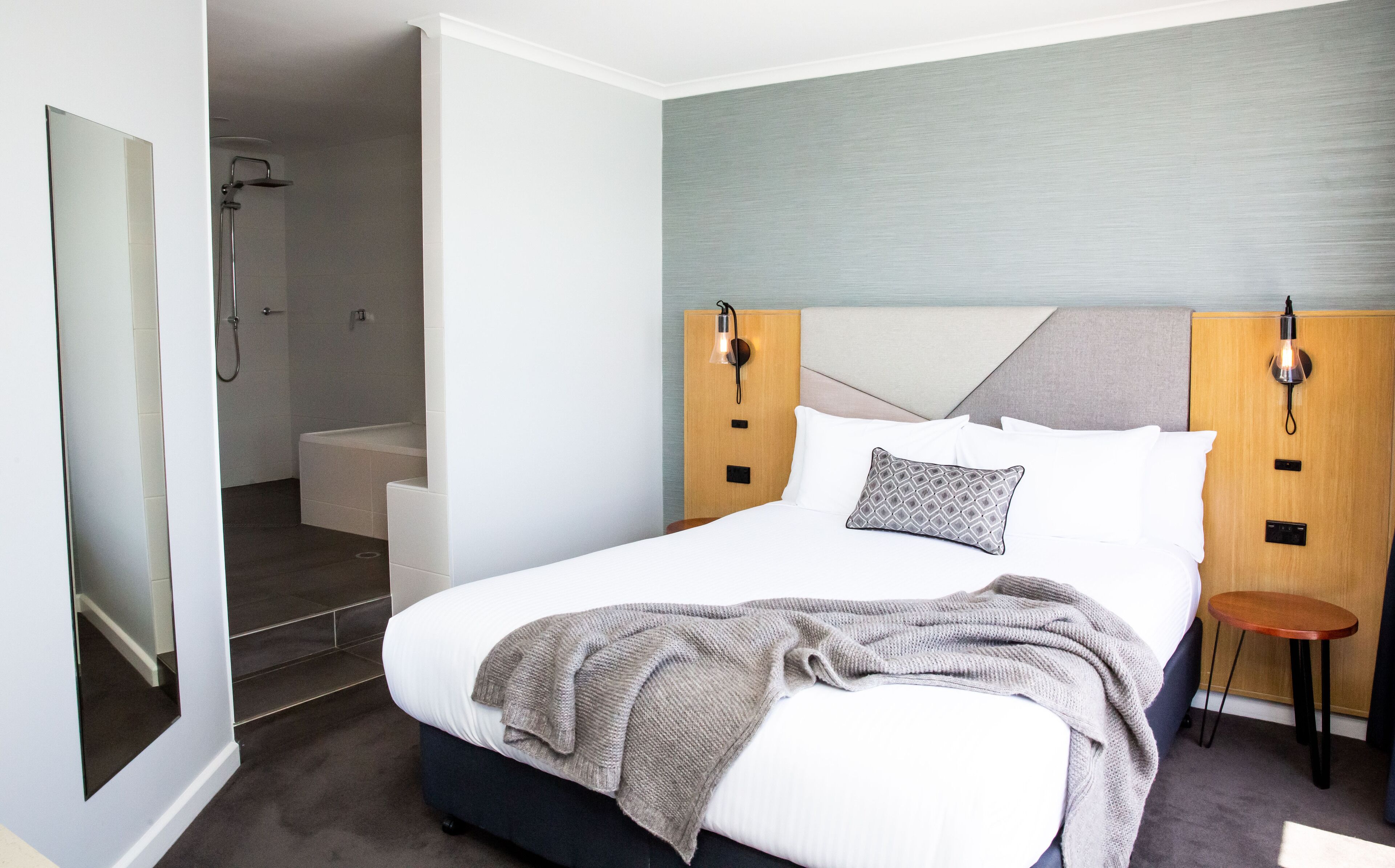 A modern hotel room with a large bed, a wooden headboard, a bedside table, and a shower area.