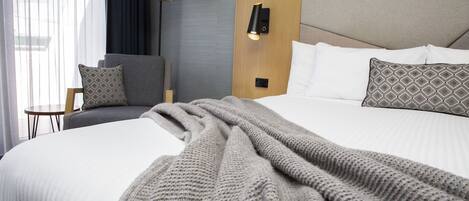 Ein ordentlich bezogenes Bett mit grauer Strickdecke, einem Holzkopfteil und einem kleinen Nachttisch mit Lampe.