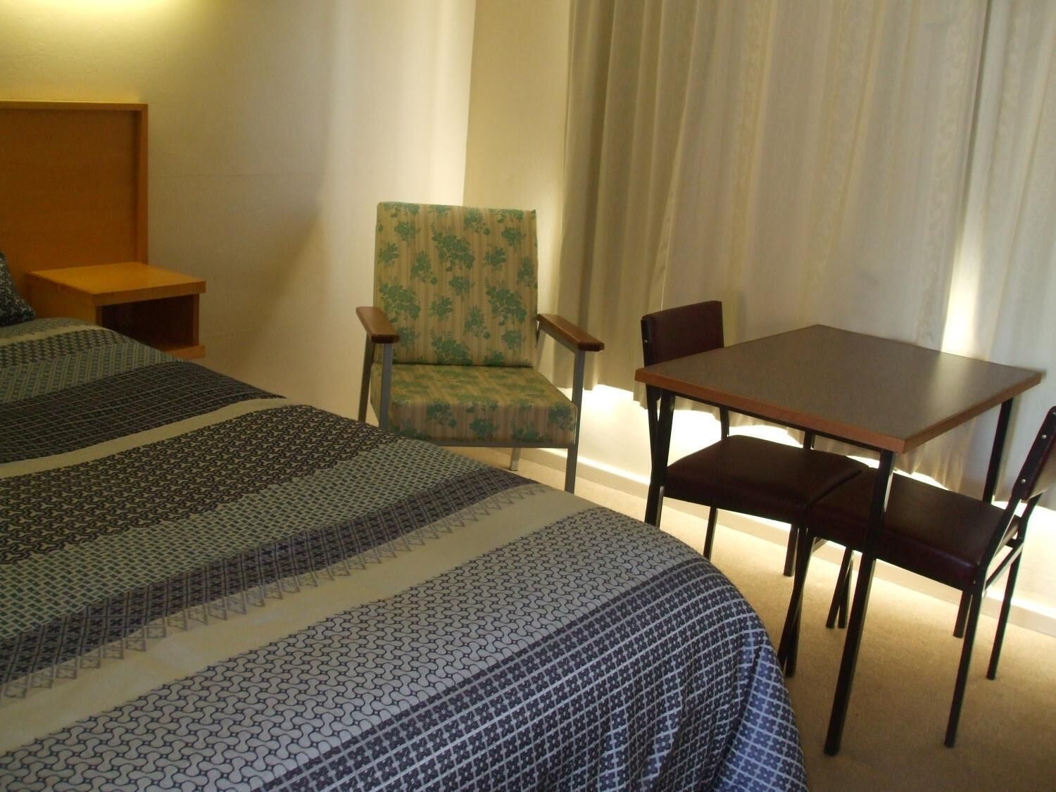 A hotel room with two beds, a wooden nightstand, a green armchair, a wooden table with four chairs, and a large window with curtains.