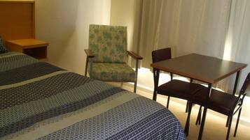 A hotel room with two beds, a wooden nightstand, a green armchair, a wooden table with four chairs, and a large window with curtains.