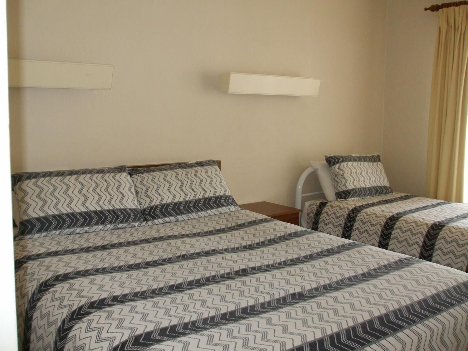 Habitación de hotel con dos camas, cada una con ropa de cama estampada. Tiene una mesita de noche y una estantería en la pared.
