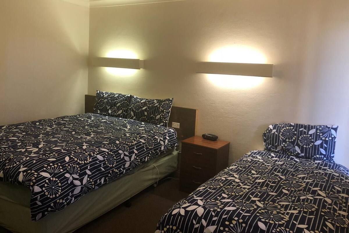 A hotel room with two beds, each with a patterned bedspread, a bedside table with a lamp, and wall-mounted lights.