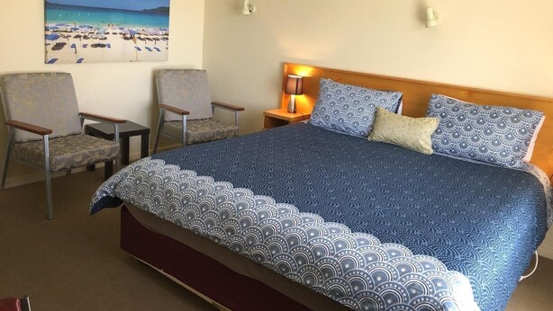 A hotel room with a large bed, two armchairs, a nightstand with a lamp, and a painting of a beach scene on the wall.
