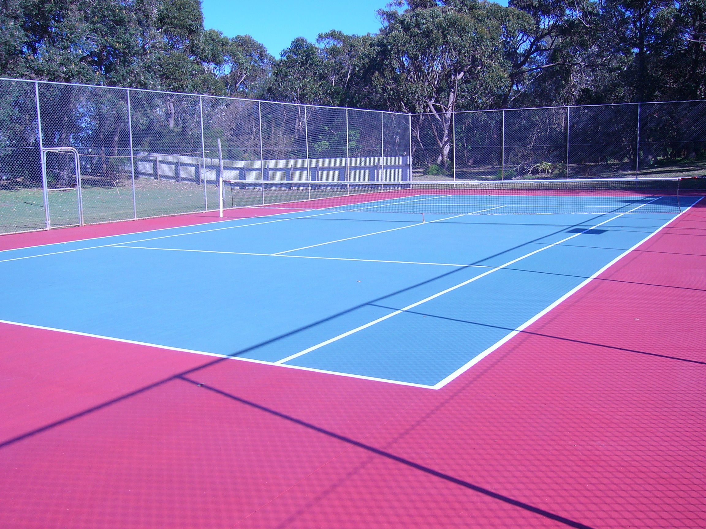 An outdoor tennis court with a red and blue surface, surrounded by a chain-link fence and trees.