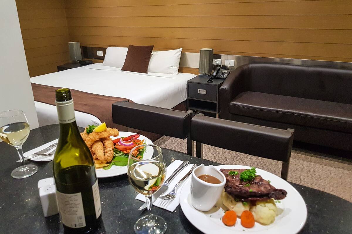 A hotel room with a bed, a sofa, a dining table with a bottle of wine, a glass, a plate of food, and a small bowl of sauce.