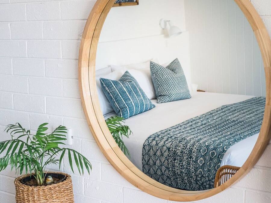 Un miroir rond fixé sur un mur de briques blanches, qui reflète un lit avec une literie et des coussins à motifs, une plante en osier et une table en bois avec une chaise.