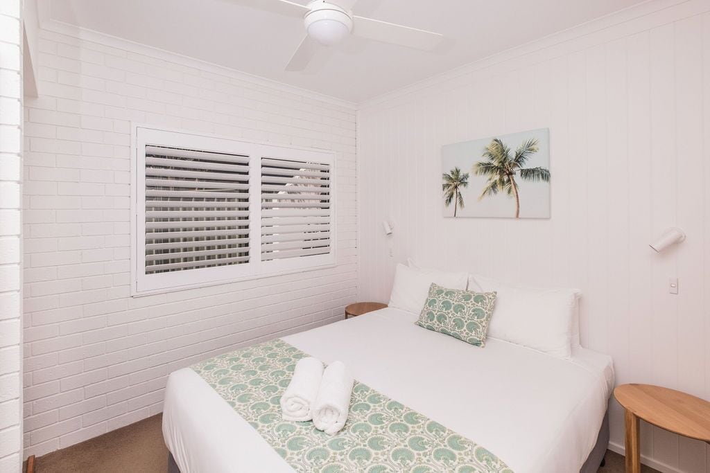 A bedroom with a bed, a window with blinds, a ceiling fan, and a wall-mounted picture of palm trees.
