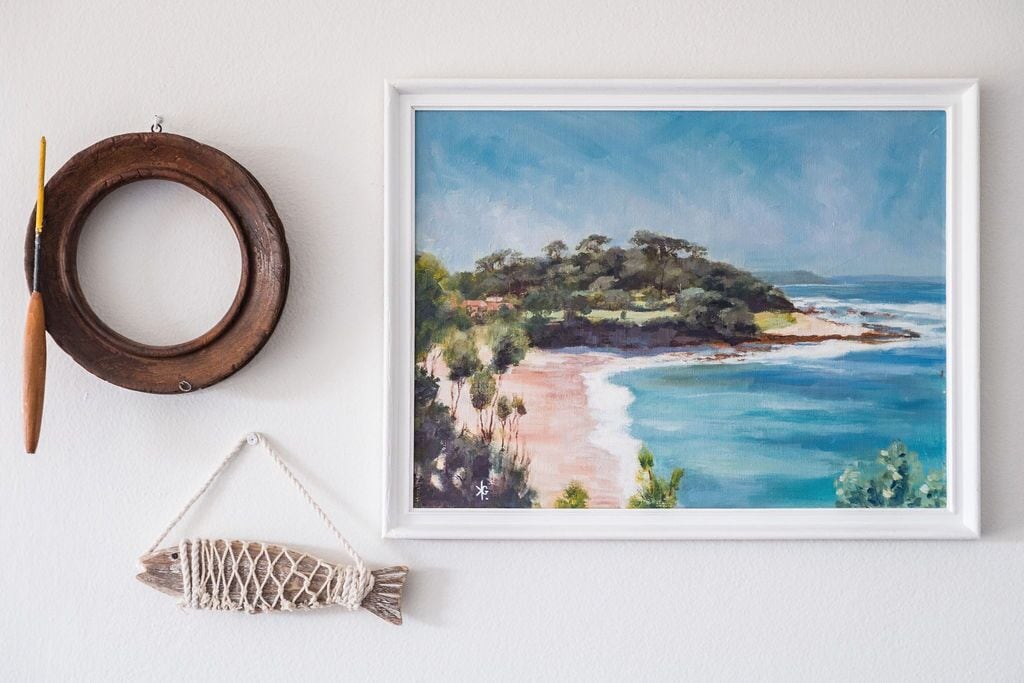 A framed coastal landscape painting, a wooden ring, and a macramé fish ornament on a white wall.
