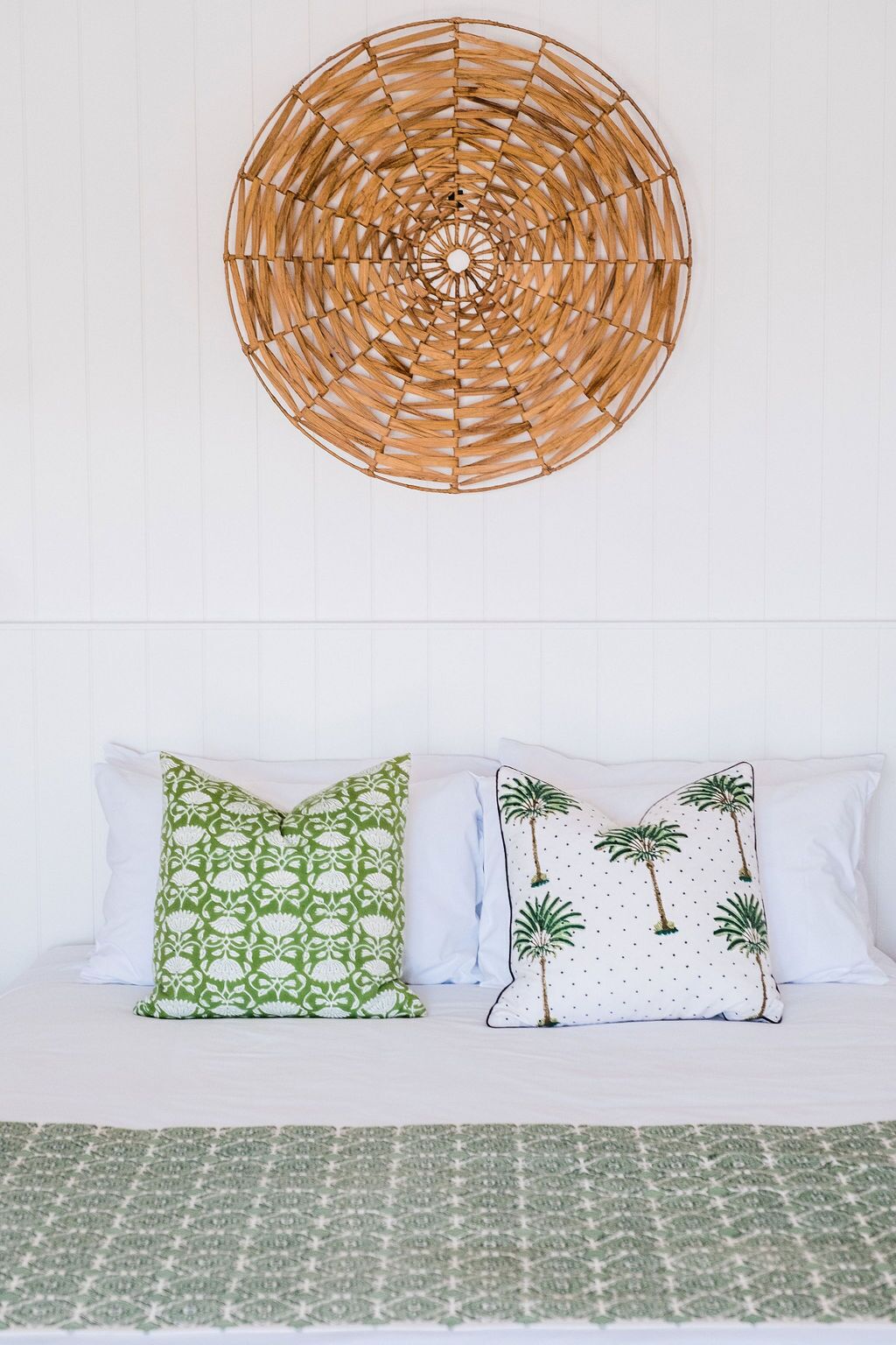 A bed with white bedding, two pillows with green and palm tree patterns, and a woven wall hanging above.