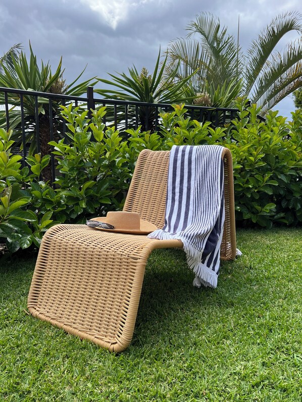 A wicker lounge chair with a striped towel, placed on a grassy lawn with lush green bushes and a palm tree in the background. - Mollymook Seascape Motel & Apartments (Ulladulla)