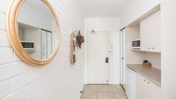 Un pasillo estrecho con pared de ladrillo blanco, un espejo con marco de madera, un microondas y un refrigerador.