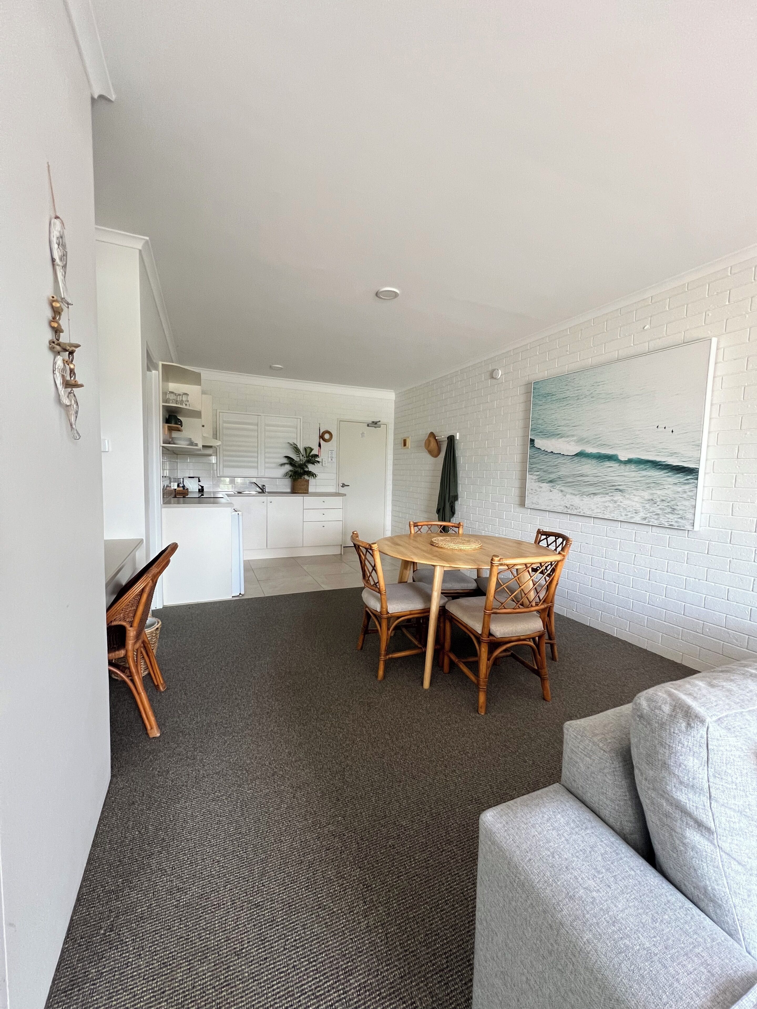 Een moderne keuken met witte kasten, een eethoek met een ronde houten tafel en rieten stoelen, en een grijs vloerkleed.