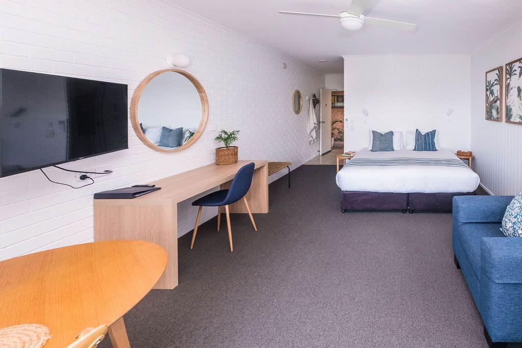 Ein modernes Hotelzimmer mit Flachbildfernseher, einem Holztisch mit Stuhl, einem Bett mit blauem Kopfteil und einem Deckenventilator.