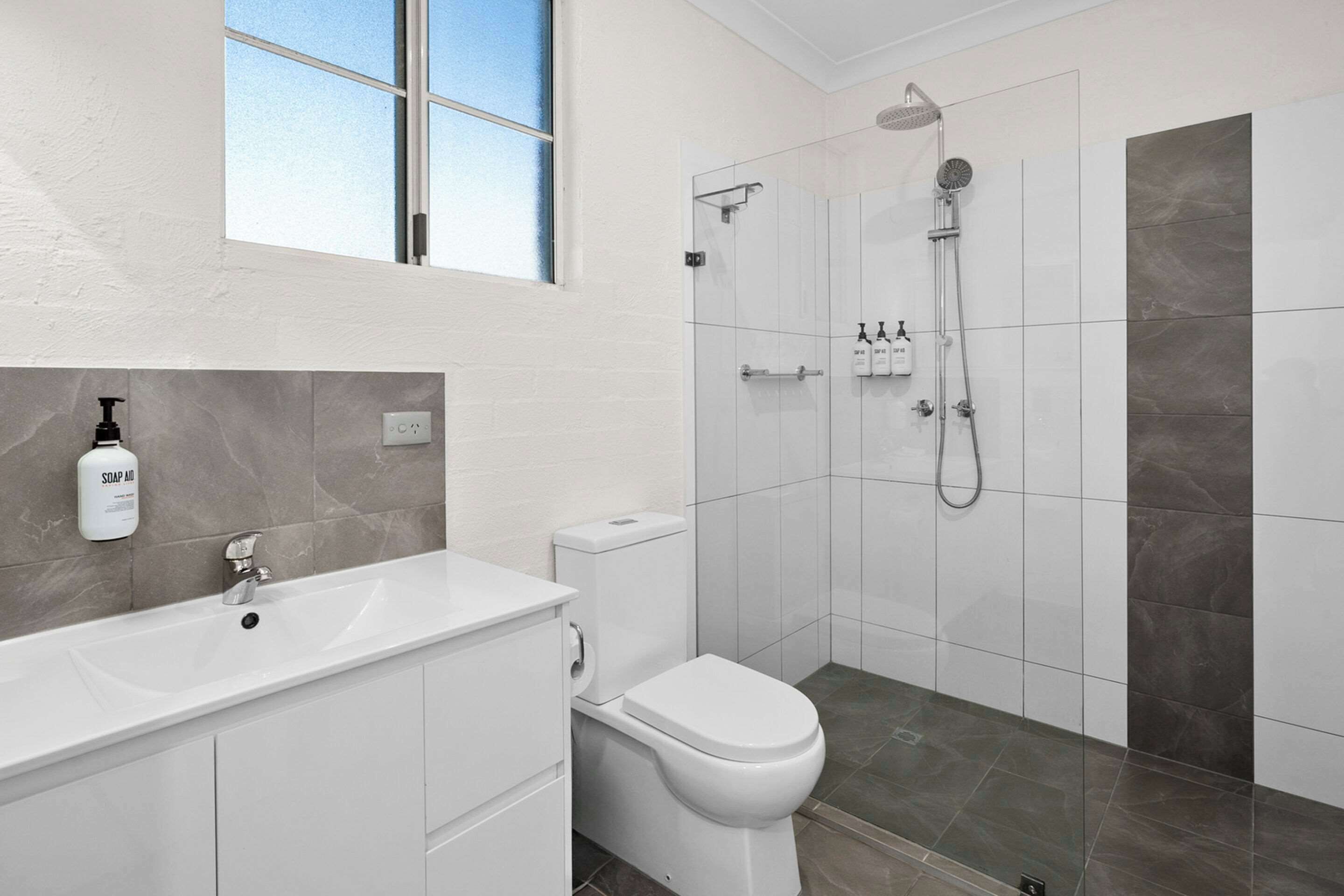 Un bany modern amb un lavabo blanc, un vàter, una dutxa amb plat de roba i una finestra sobre el lavabo.