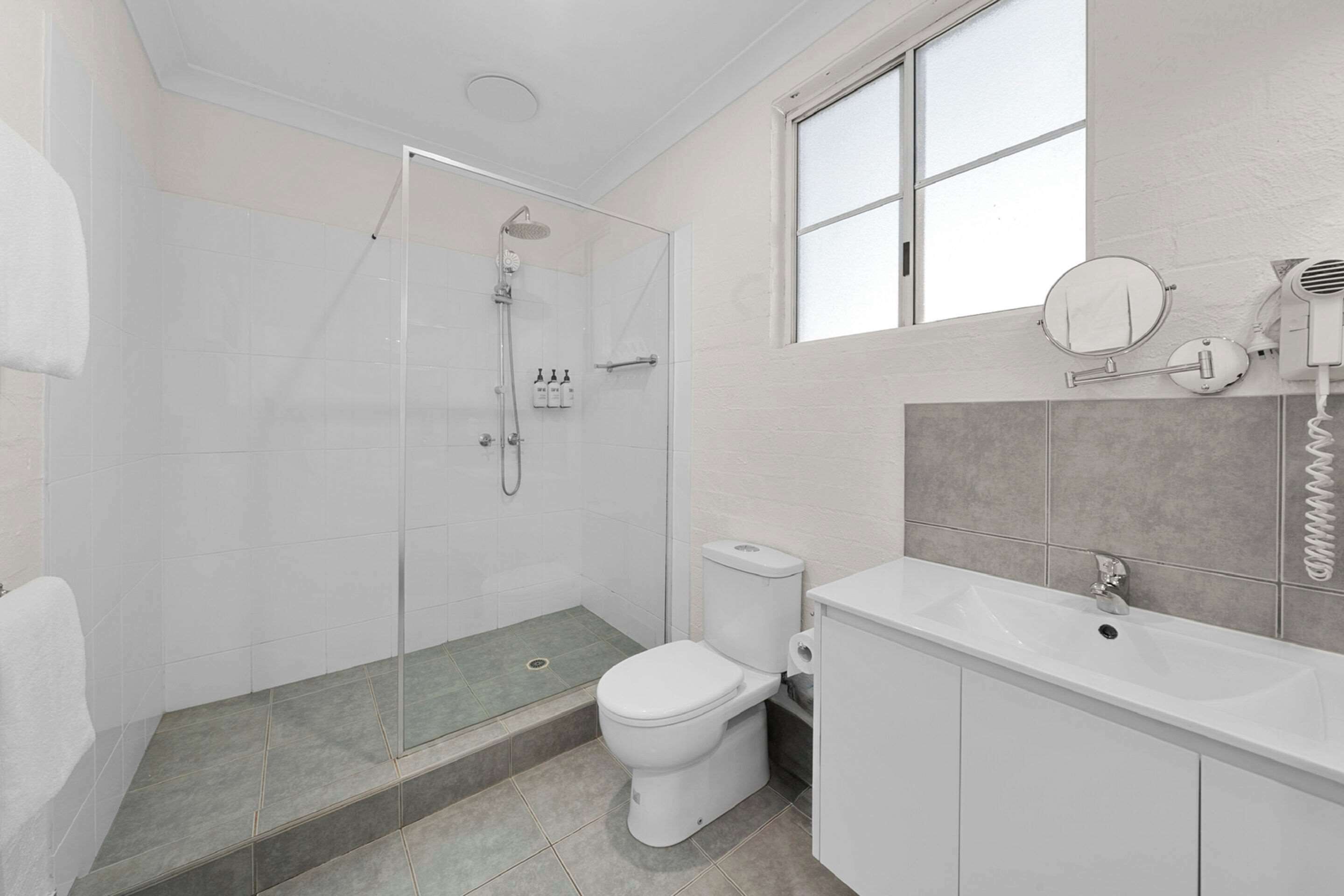 Uma casa de banho moderna com cabine de duche em vidro, sanita branca e toucador com espelho e secador de cabelo.