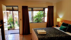 A hotel room with a bed, a nightstand, a lamp, and a view of greenery outside. - Rockhampton Palms Motor Inn (Rockhampton)