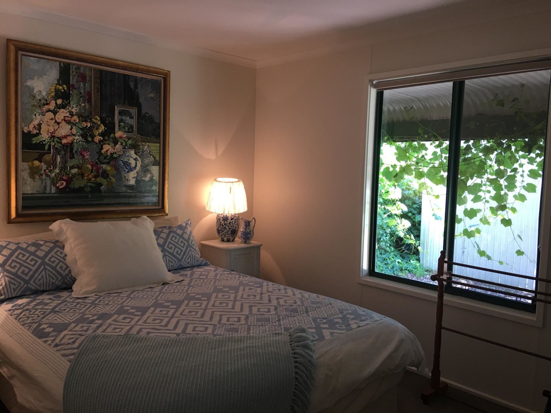 Un dormitorio con cama, mesita de noche con lámpara, un cuadro y ventana con vista a follaje.