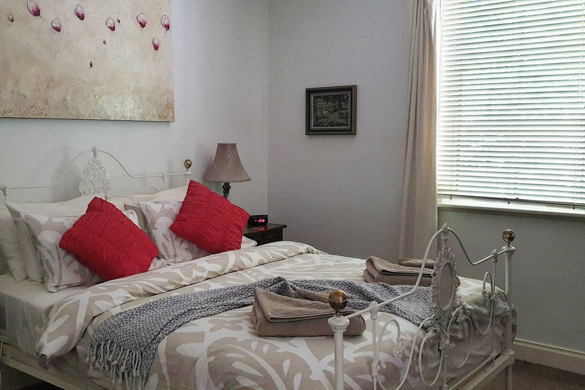 A bedroom with a bed, two bedside tables, a nightstand, a lamp, a blanket, and a window with blinds.