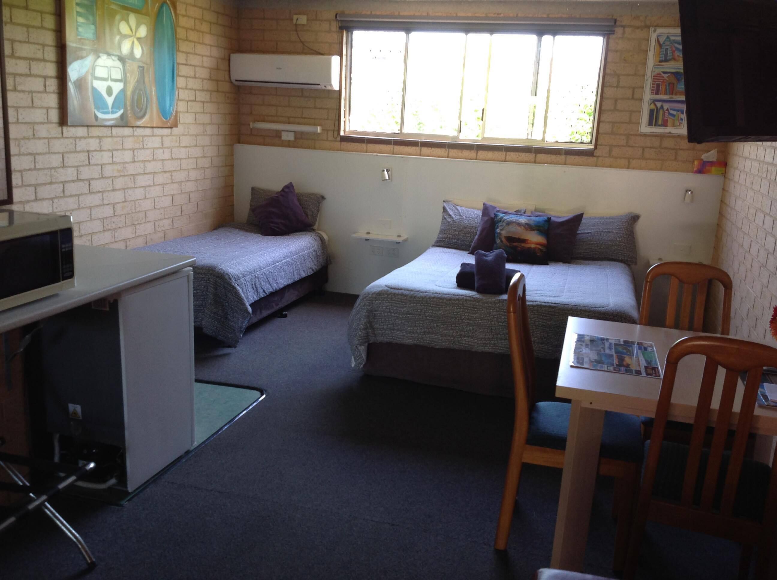 A dormitory room with two single beds, a desk with a chair, a microwave, and a window with blinds.