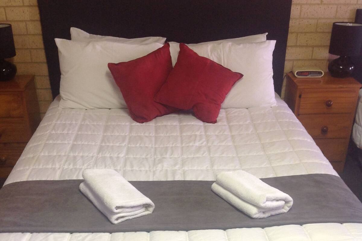 Een keurig opgemaakt bed met witte en rode kussens, een grijze deken en twee opgevouwen handdoeken.