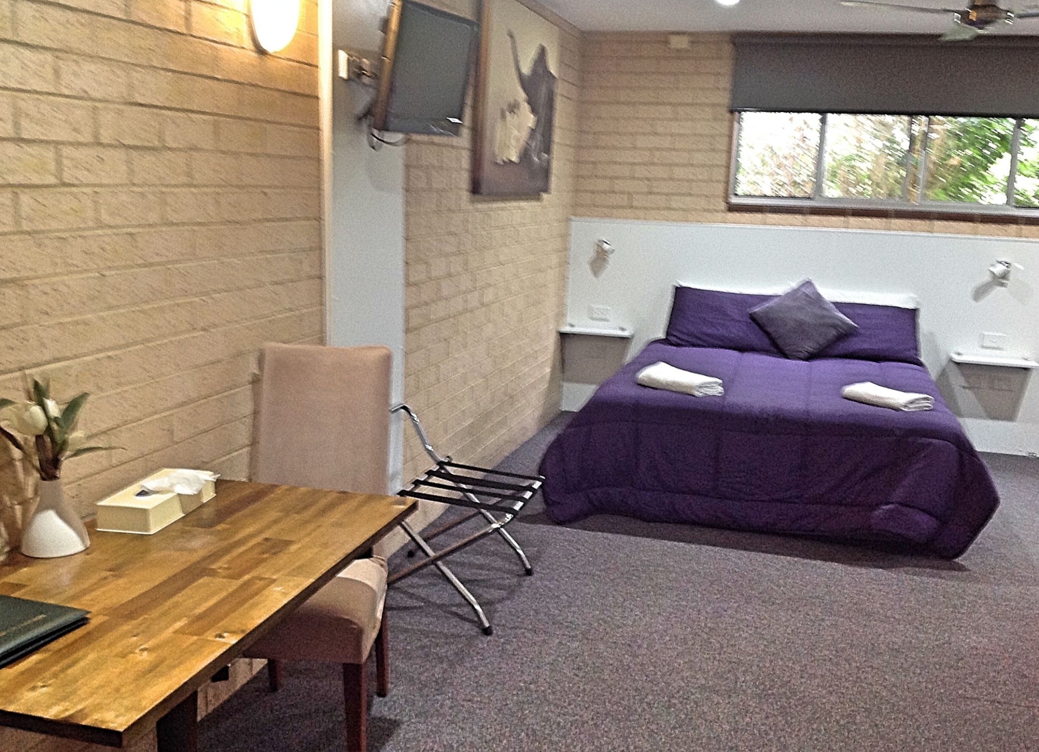 A room with a wooden table, a chair, a bed with a purple bedspread, and a window with a view of trees.