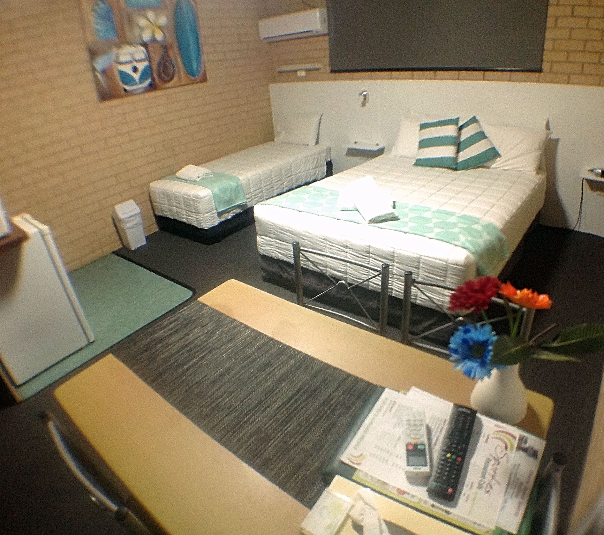 A small hotel room with two single beds, a desk with a vase of flowers, and a TV on the wall.