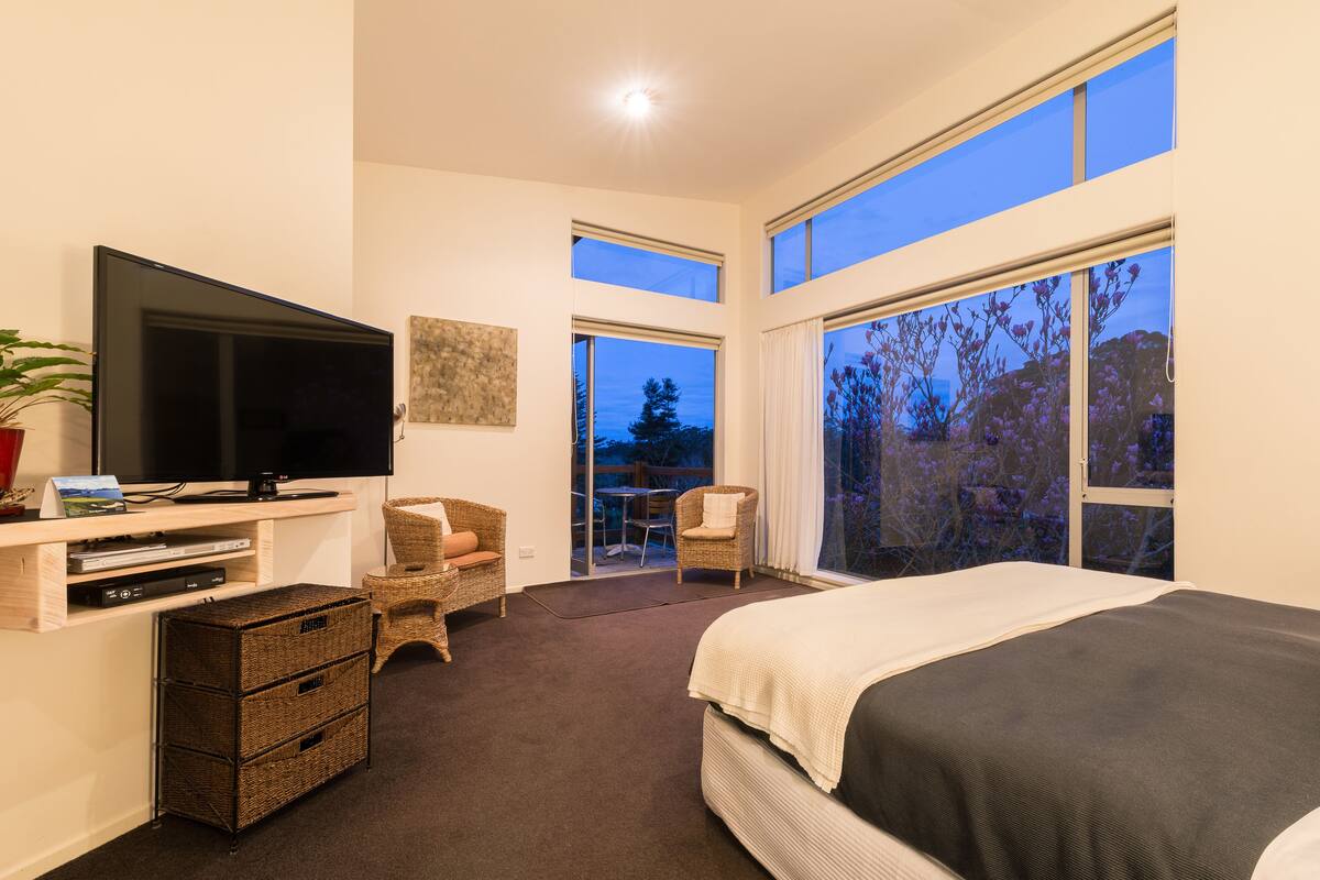 A bedroom with a large bed, a TV, a desk with a computer, and a balcony with a view of trees.