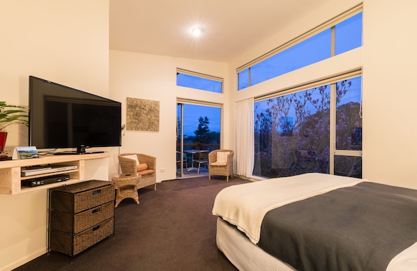 A bedroom with a large bed, a TV, a desk with a computer, and a balcony with a view of trees. - Stone Store Lodge (Kerikeri)