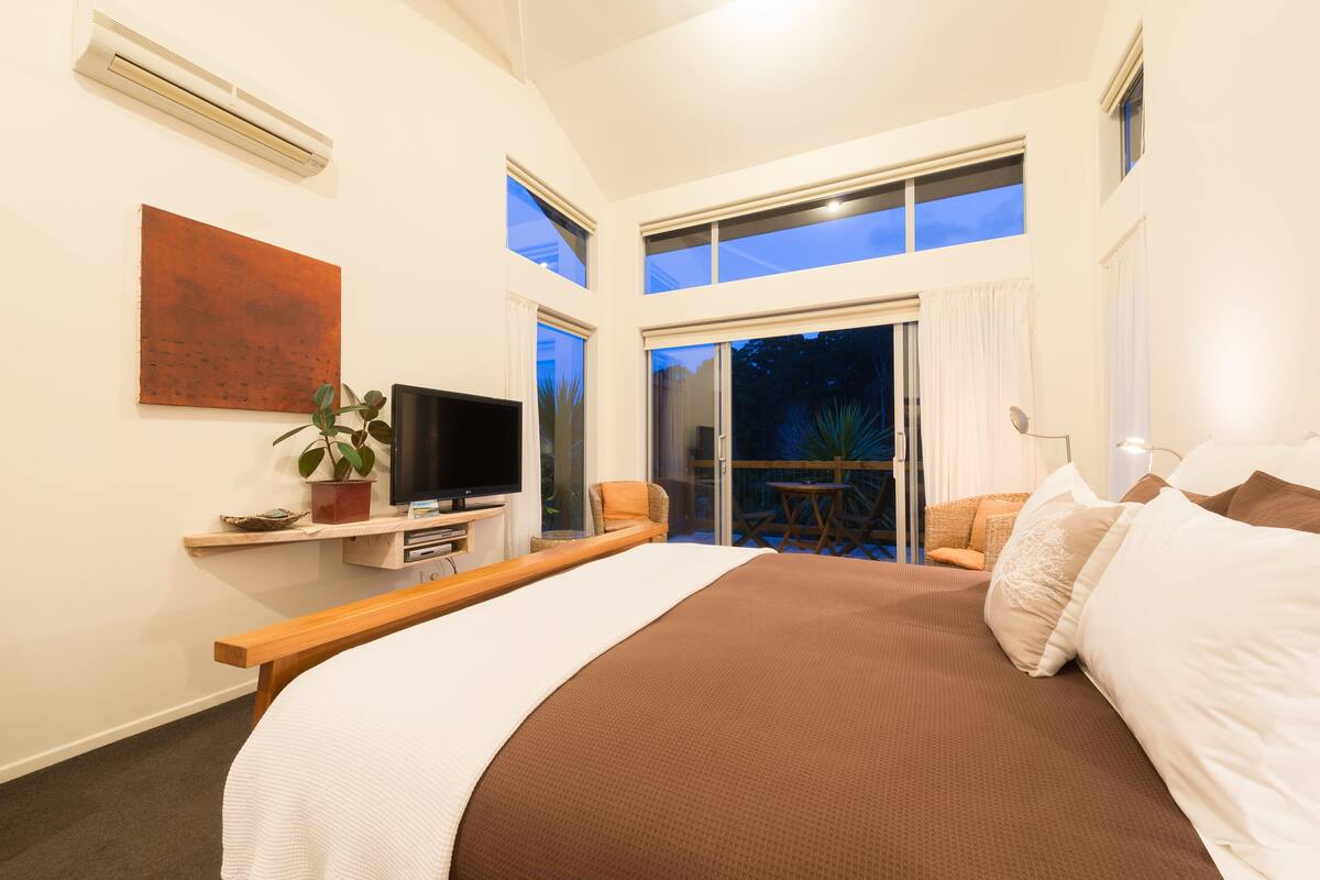 A modern bedroom with a large bed, a flat-screen TV, a plant, and a sliding glass door leading to a balcony.