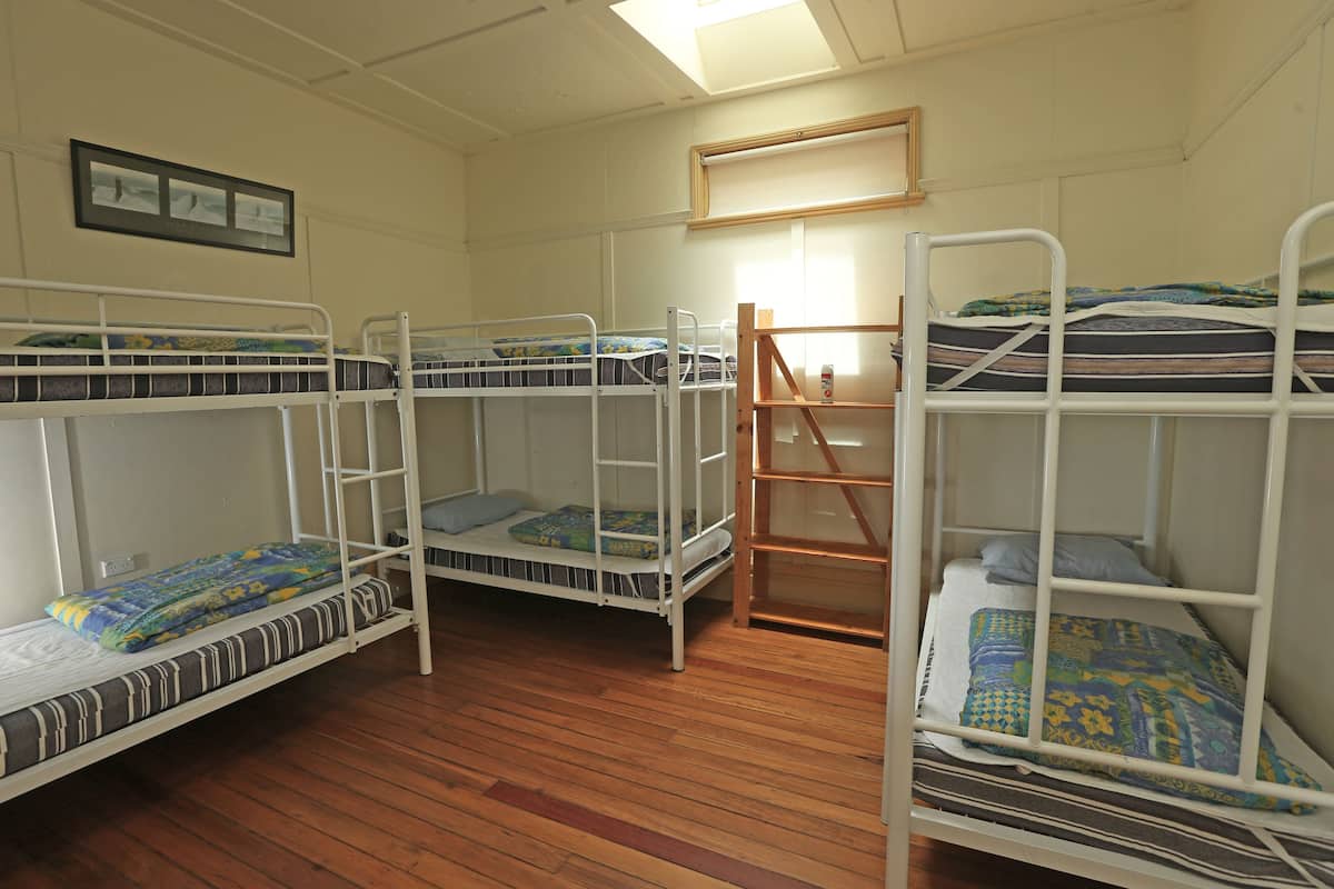 A dormitory room with three bunk beds, wooden flooring, and a window with blinds.
