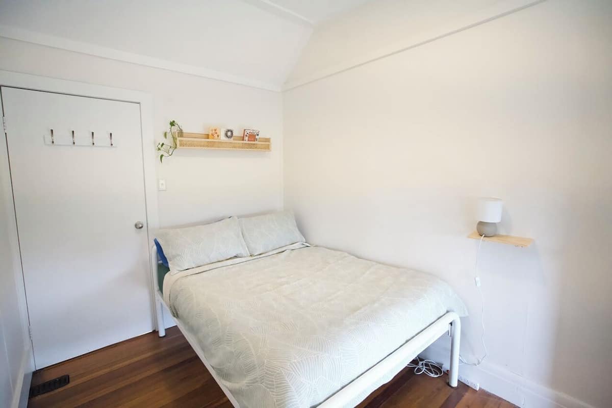 A bedroom with a bed, a door with hooks, a shelf with decorative items, and a bedside table with a lamp.