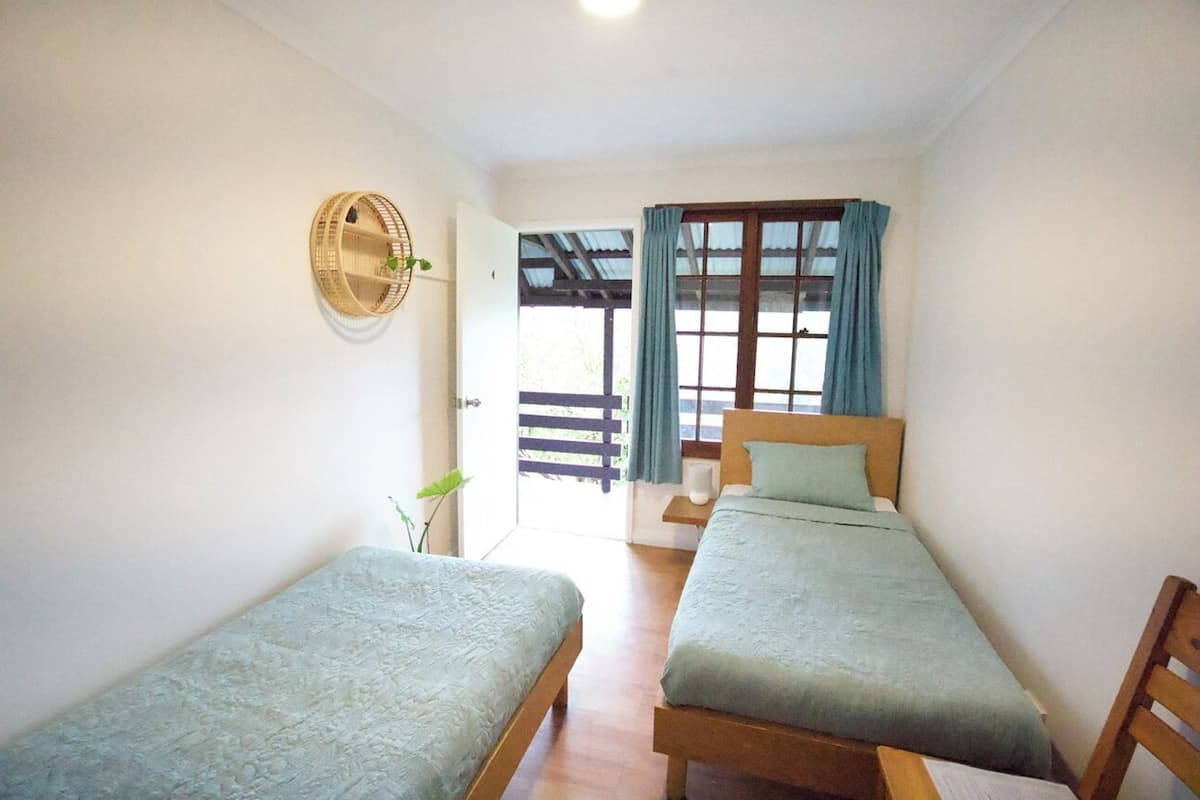 A room with two single beds, a wooden headboard, a window with curtains, and a wall-mounted fan.