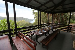 Un espacio exterior cubierto para comer con bancos y mesas de madera, con vistas panorámicas a un lago y colinas.