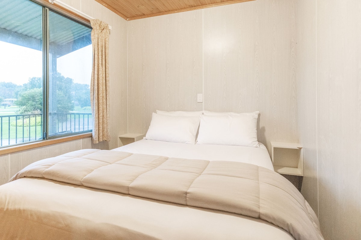 A bedroom with a large bed, two bedside tables, a window with a curtain, and a view of greenery outside.