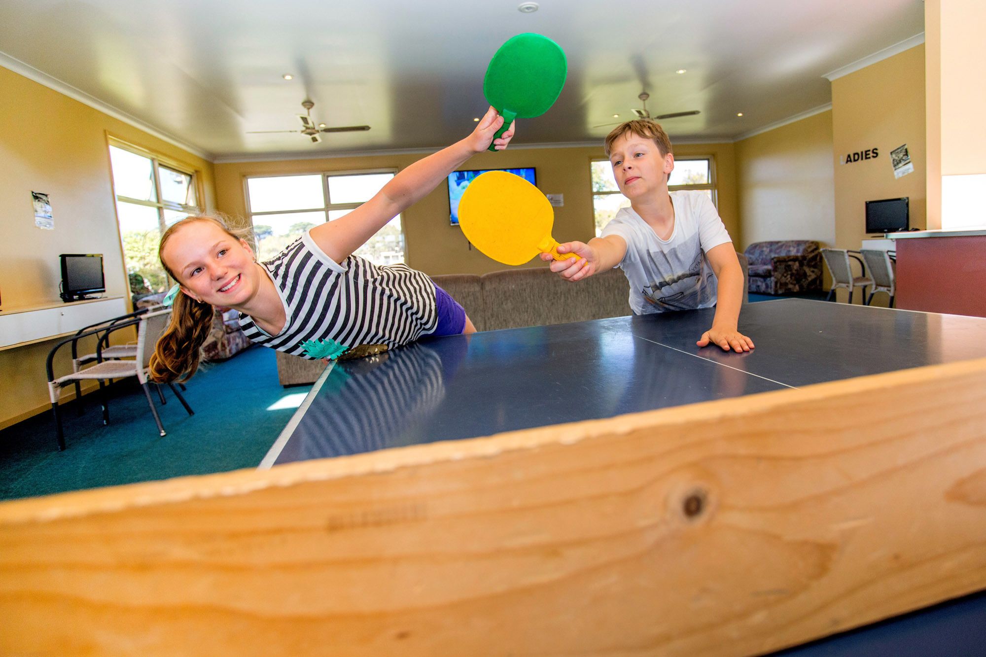 Duas pessoas jogando tênis de mesa em um salão com uma mesa de ping pong, cadeiras e uma televisão ao fundo.